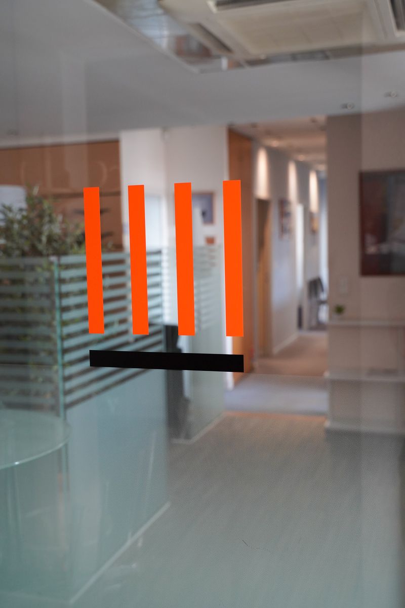Le logo du cabinet, quatre colonnes orange sur un socle noir, collé sur la porte vitrée qui donne sur le couloir